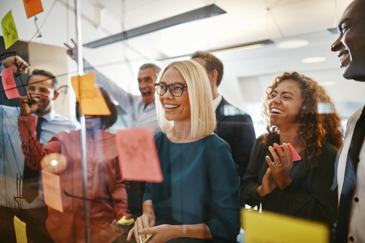 A diverse team smiling at a transparent white board with colorful sticky notes and writing on it